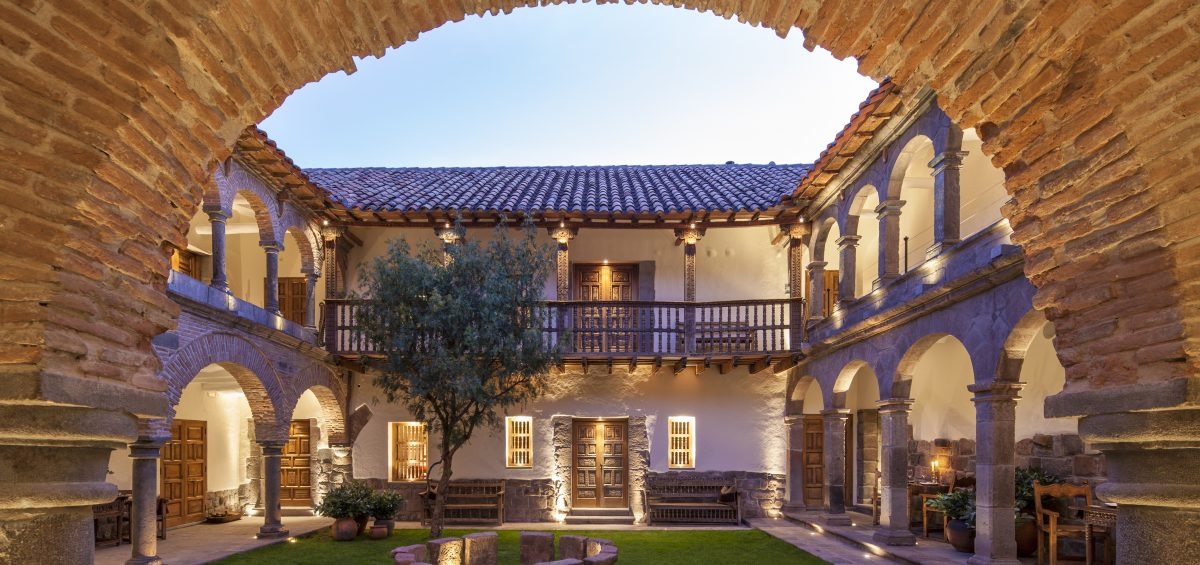 Two-storey stone courtyard at dusk with arched colonnades, a carved balcony and a single olive tree on the lawn