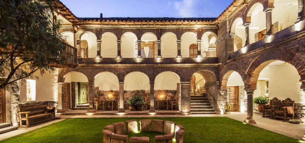 Colonial courtyard illuminated at night, with stone archways, a carved wooden staircase and central stone hearth