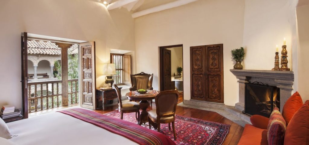 Suite with carved wooden doors opening onto a colonnaded balcony, a stone fireplace and striped Andean bed runner