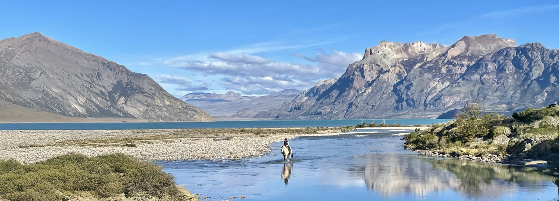 Horse and rider crossing a shallow river with dramatic Patagonian mountains rising in the background.