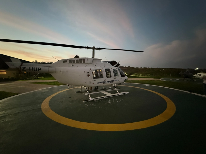 A helicopter rests on a floodlit helipad at dusk, ready for a scenic flight across the Zambezi region as evening light fades. 