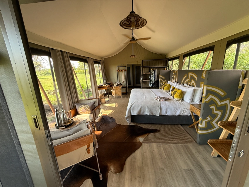 A light-filled safari tent reveals a comfortable bedroom with canvas walls and open views across the surrounding wilderness. 
