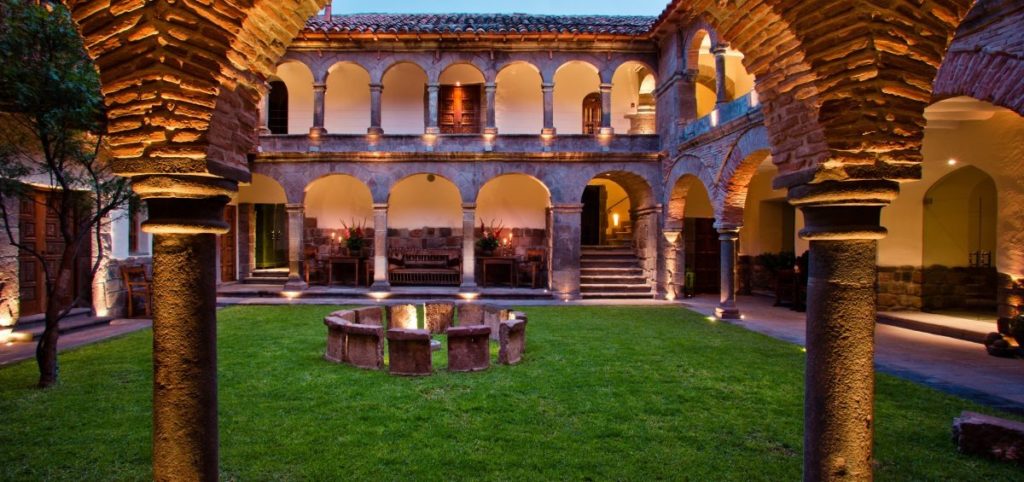 Illuminated colonial courtyard at dusk with a stone seating circle on the lawn and arched galleries above