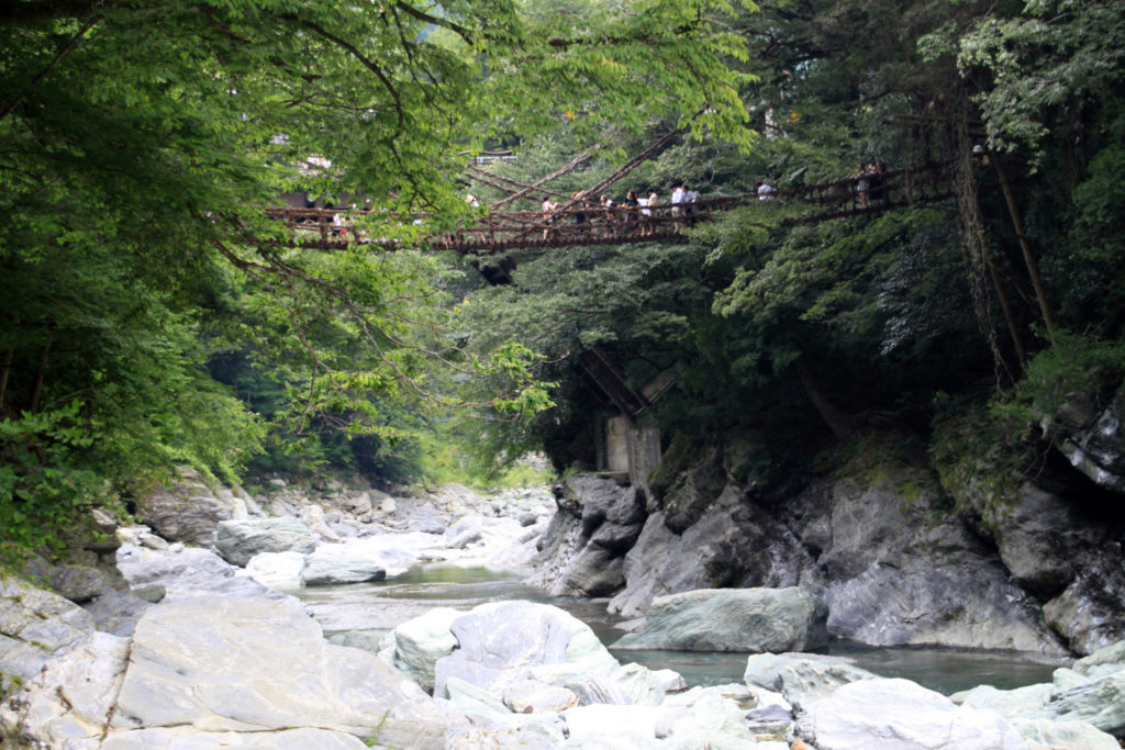 Visitors cross a bridge woven from mountain vines, high above a clear green river and pale boulders, in a deep forested gorge in Japan’s Iya Valley.
