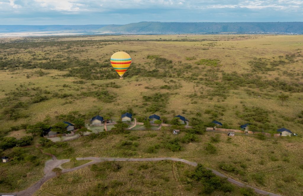 Aerial view of Kandili Camp's tents in a line along acacia scrub, with a striped hot air balloon floating over the plains and an escarpment beyond.