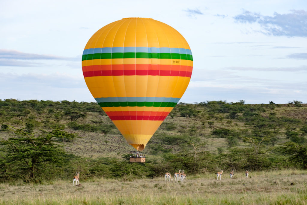 Striped hot air balloon drifting low over acacia scrub as Thomson's gazelles graze in the grass below, near Kandili Camp, Kenya.
