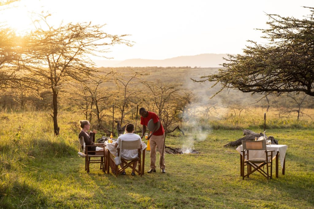 Guests dining outdoors at Kandili Camp in golden evening light, a staff member serving drinks beside a smouldering campfire under acacias.