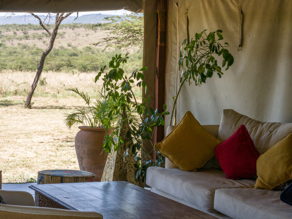Kandili Camp lounge with a cream sofa, yellow and red cushions and a terracotta planter, open to acacia-dotted plains and distant hills.