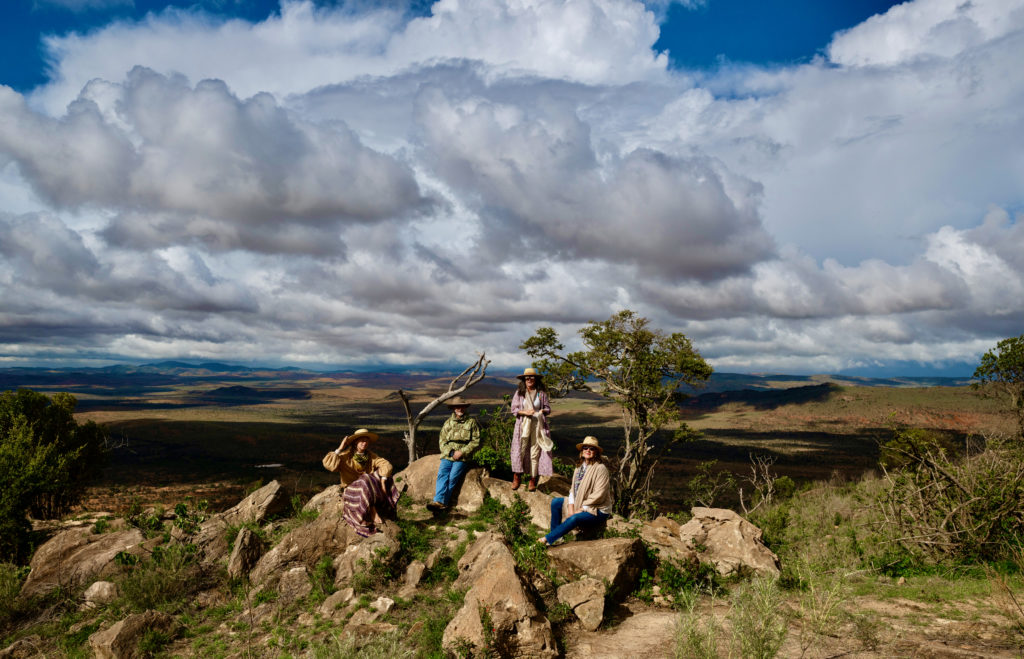 Four guests in wide-brimmed hats rest on a rocky outcrop in the Lolldaiga Hills, with storm clouds building over the Laikipia plains.