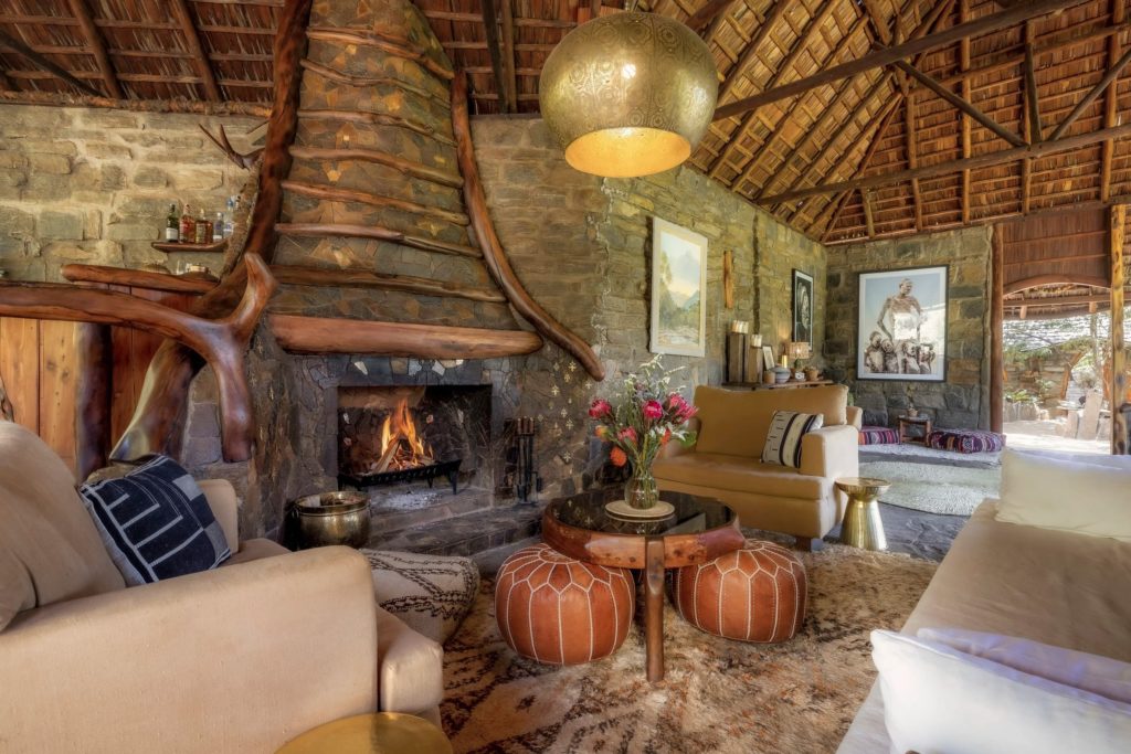 Main lounge at Desert Rose with a stone fireplace, carved wooden beams, a thatched roof, leather poufs and a brass pendant lamp.