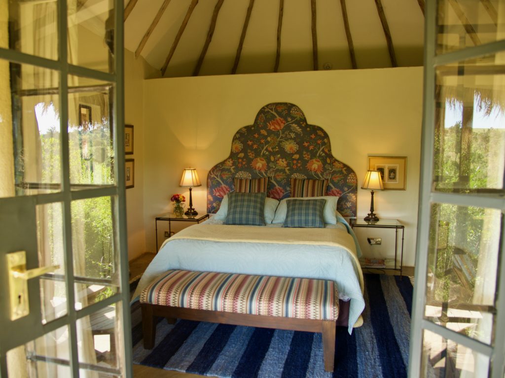 Round bedroom seen through open glass doors, with a floral upholstered headboard, plaid cushions, a striped bench and a blue striped rug.