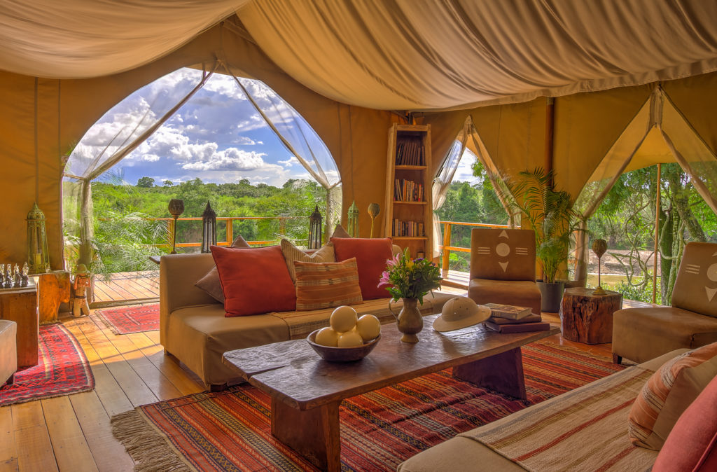 Naibor's main lounge tent is furnished with cushioned sofas, a bookshelf and safari curios, opening on two sides to views of the surrounding woodland.