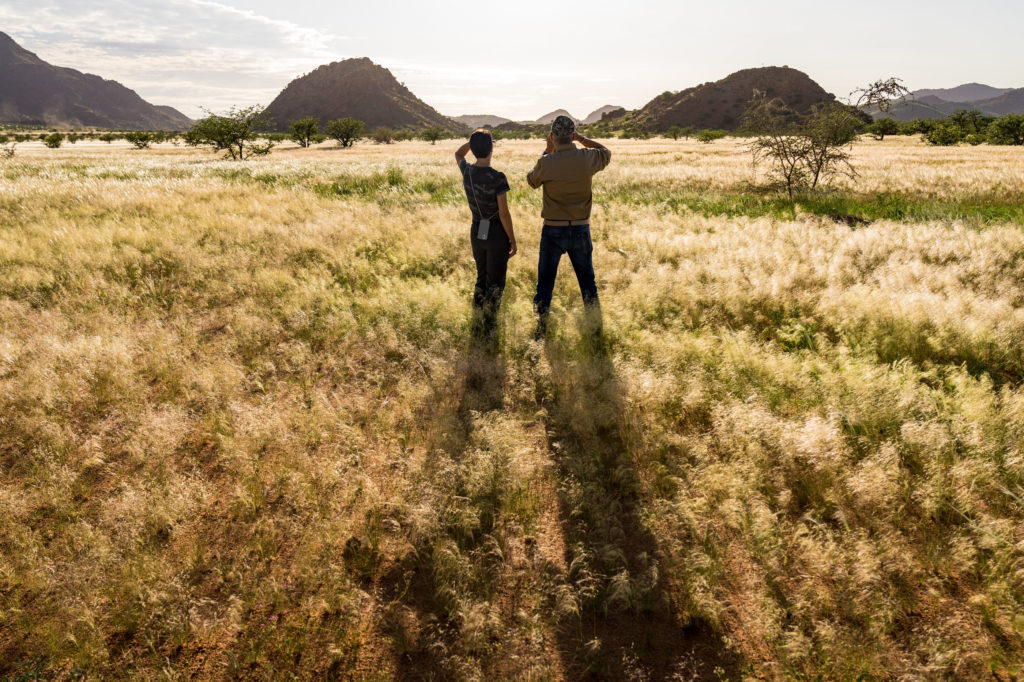 Two guests pause on a self-guided walking trail, scanning a green Damaraland valley scattered with trees and distant hills.