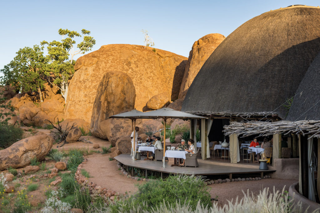 Guests breakfast beneath umbrellas on Mowani's boulder-bound dining deck, thatched roofs and sunlit rock rising close behind them.
