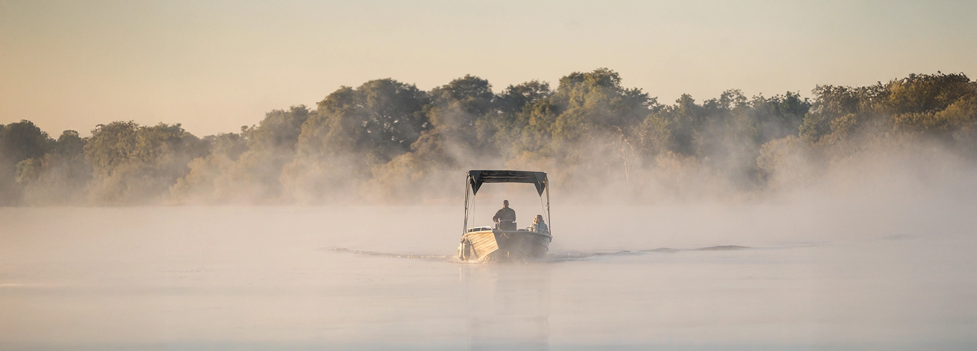 A small boat glides through early morning mist on the Zambezi River, capturing the quiet, atmospheric beauty of a river safari near Mukwa River Lodge.
