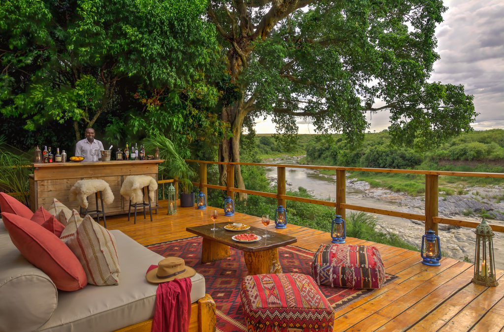 A bartender stands ready at Naibor's riverside bar deck, furnished with sheepskin stools, cushioned seating and lit lanterns overlooking the river below.