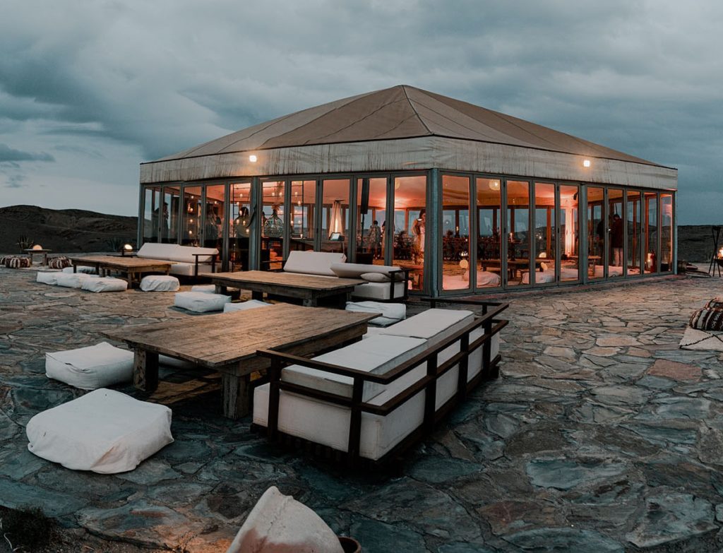 Dusk settles over a glass-walled dining pavilion, its warm interior lights spilling onto cushioned outdoor seating.