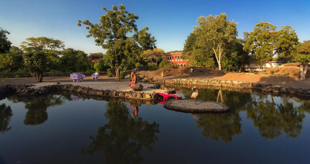 A guest relaxes by a lily pond at Orchard Palace, Gondal, with a stork on a small island and the palace visible beyond the trees