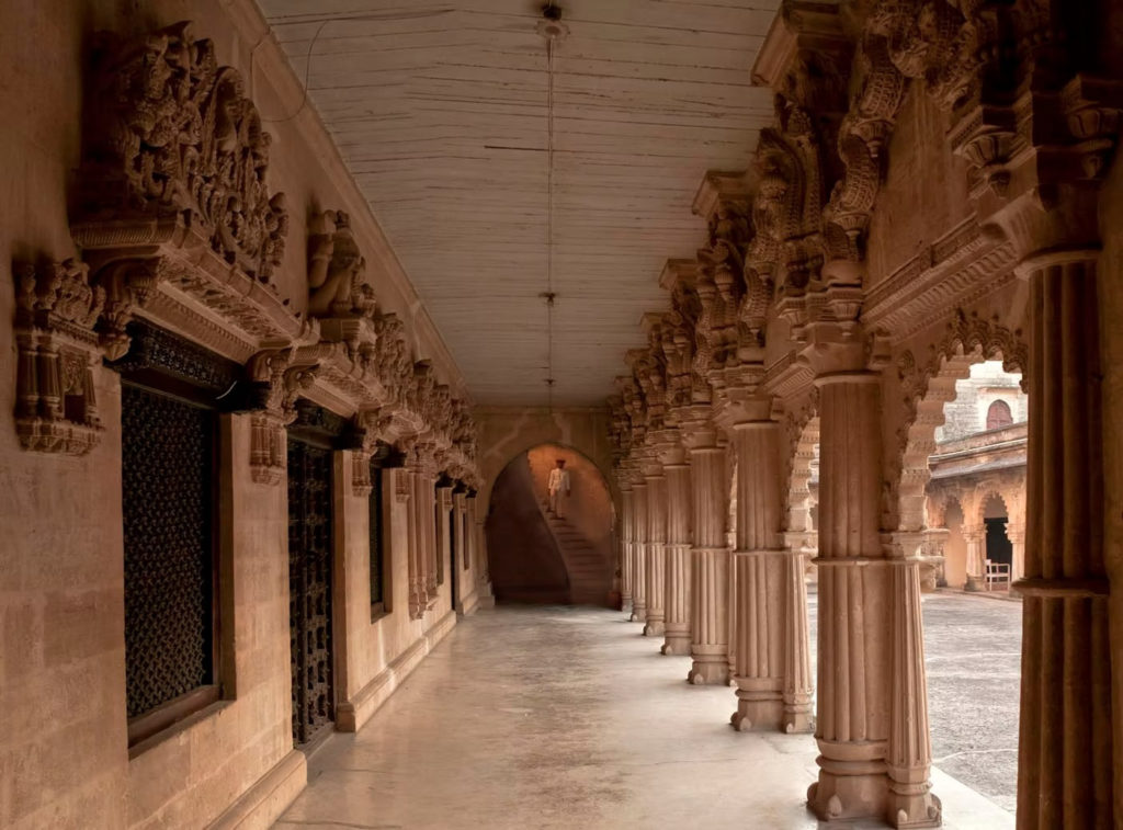 A colonnaded stone corridor with carved pillars leads through the historic Huzoor Palace complex near Orchard Palace, Gondal