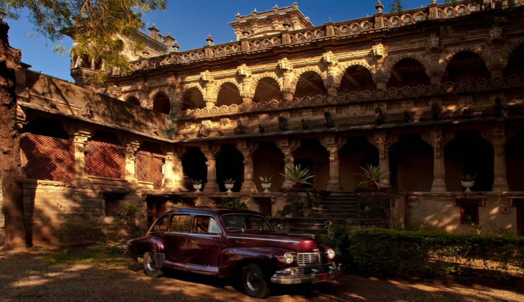 A vintage car rests in a sunlit courtyard beneath the elaborately carved stonework of the Gondal palace complex