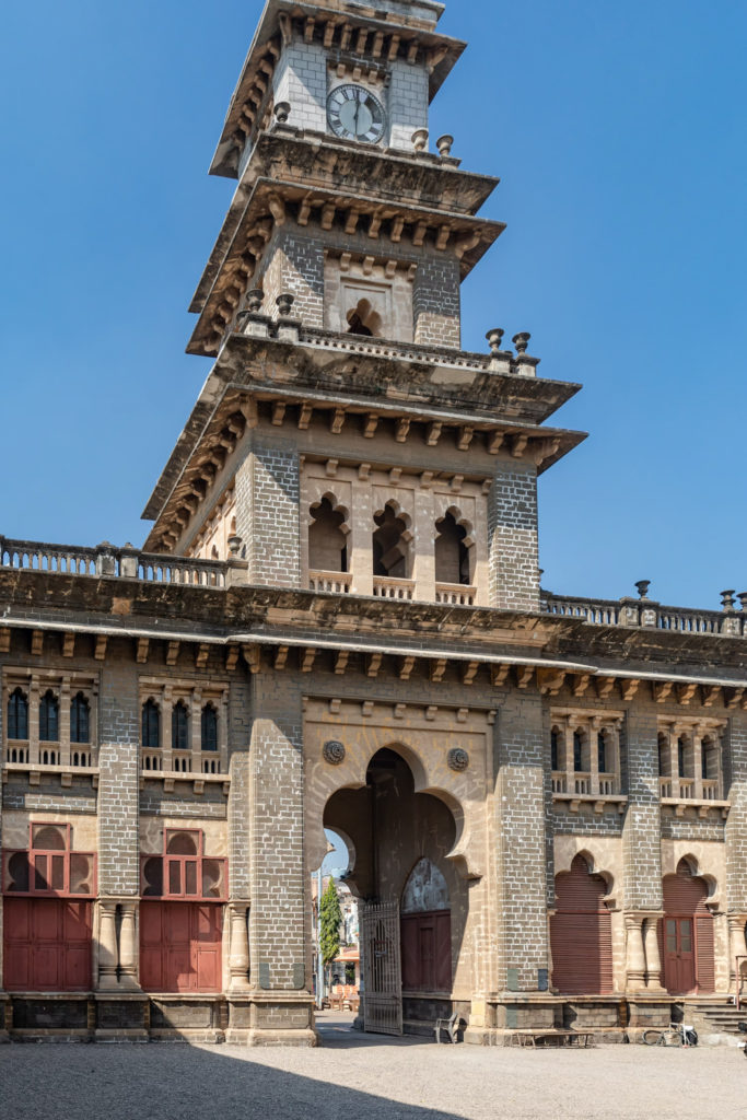 The clock tower of Huzoor Palace rises above arched stone windows near Orchard Palace, Gondal
