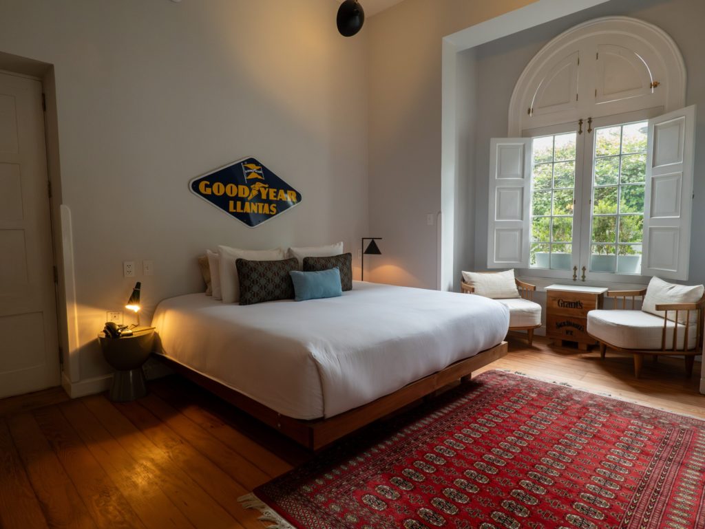Bedroom with a vintage Goodyear enamel sign above the bed and an arched window opening onto a leafy courtyard
