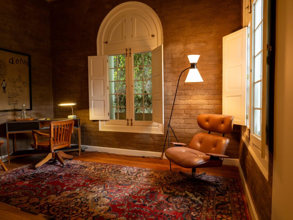 Exposed-brick sitting room with a tan leather lounge chair, floor lamp and vintage desk beside an arched shuttered window