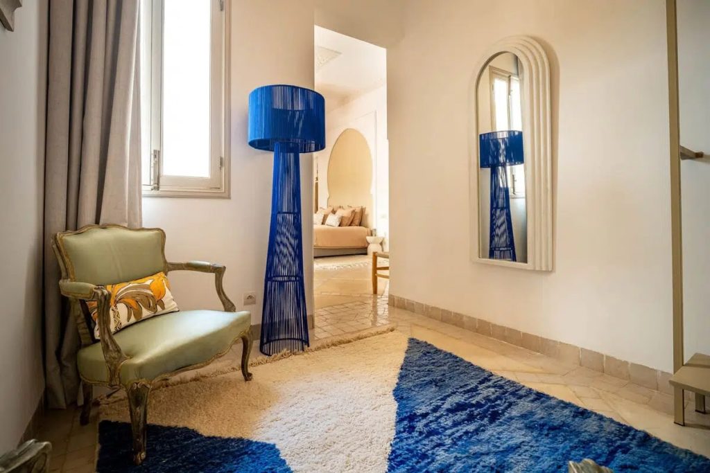 Corridor at Riad Malaika with sage armchair, arched mirror & blue rattan floor lamp