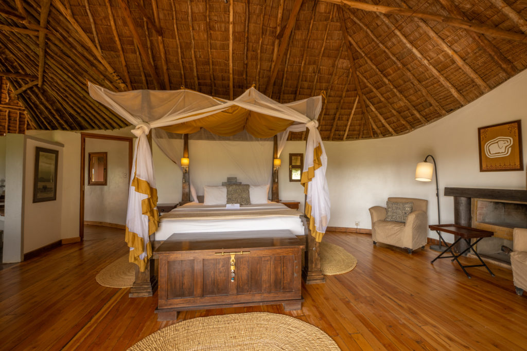 Bedroom at Tawi Lodge with a four-poster bed under a mosquito net, wooden trunk, fireplace and high thatched ceiling.