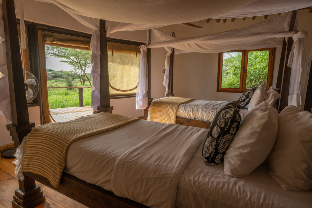 Twin room at Tawi Lodge with two four-poster beds under mosquito nets and an open canvas door onto a deck facing acacia bush.