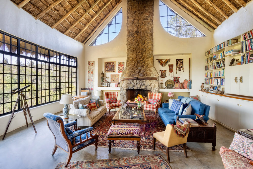 Sitting room at Olepangi Farm, Kenya, with a stone fireplace, blue sofa, checked wing chairs, a brass telescope and a tall thatched ceiling.