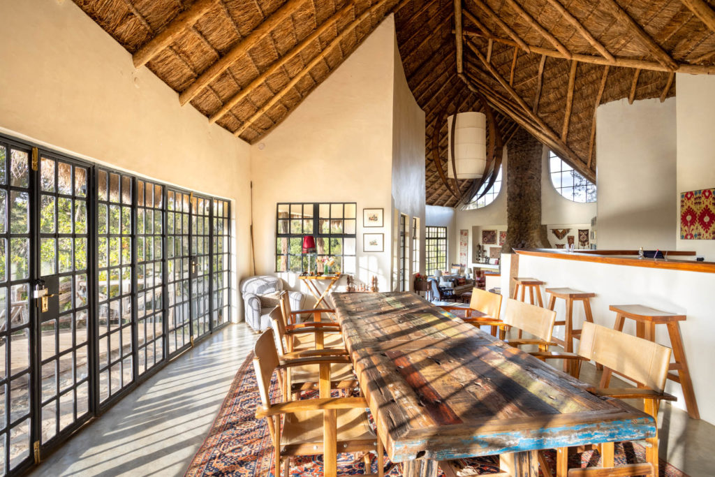 Long weathered-wood dining table with canvas chairs under a high thatched roof, late sun striping the floor through steel-framed doors.