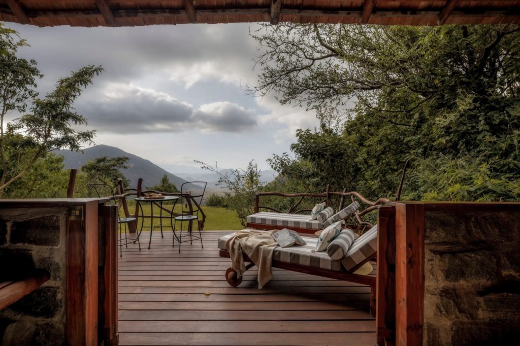 View from the bed across a wooden veranda with striped loungers and a bistro table, looking out to layered hills under a grey sky.