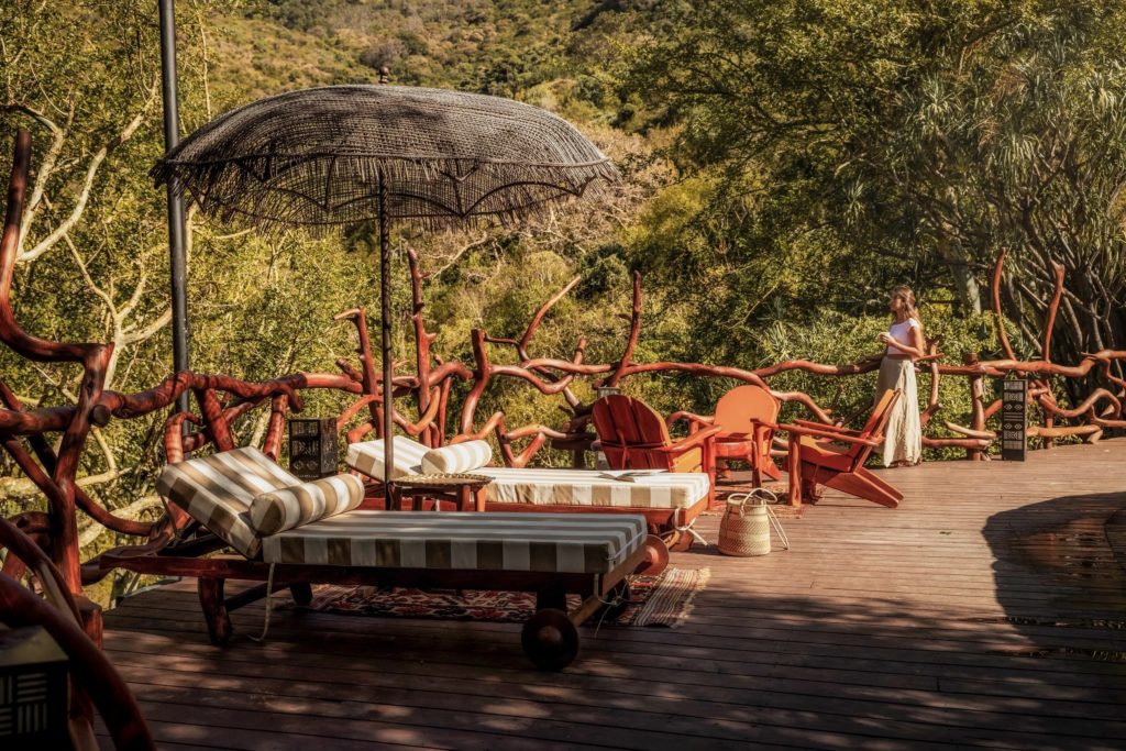 A guest stands at a branch-work railing on the pool deck, beside striped loungers under a woven parasol in dappled sunlight.