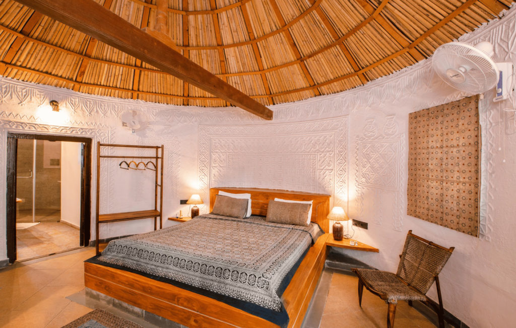 Kooba cottage bedroom by lamplight, with a carved bamboo ceiling and patterned bedspread against textured white walls.