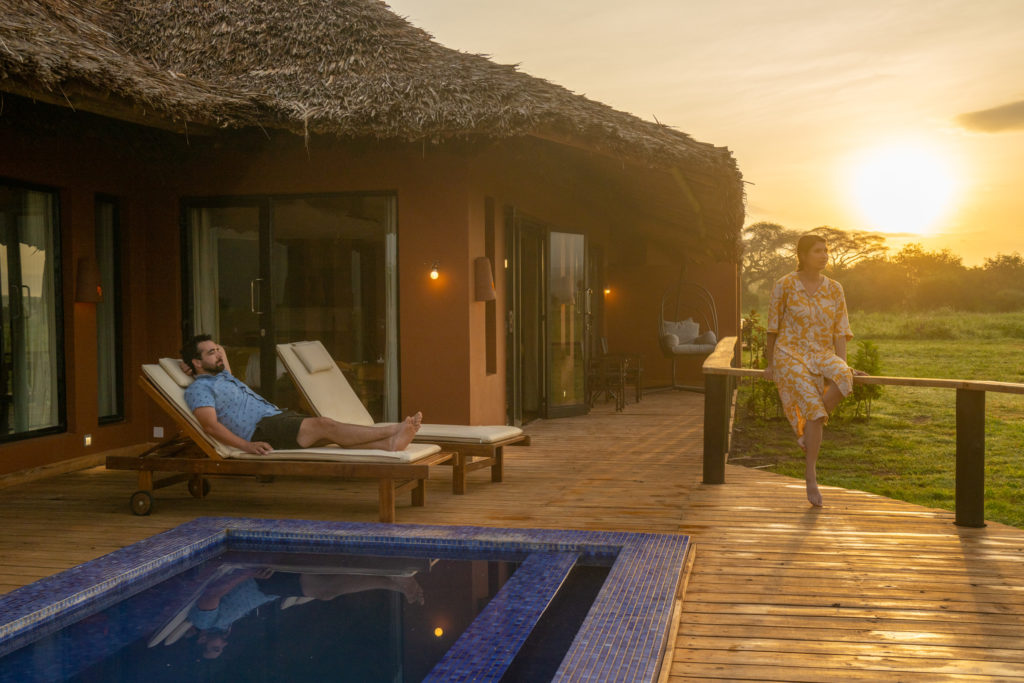 Two guests relax on a suite veranda beside a mosaic plunge pool at sunrise, one on a lounger and one perched on the deck rail.