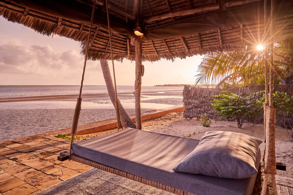 Cushioned swing daybed under a thatched roof facing the sea and tidal sand flats, with low sun glowing through palm fronds.