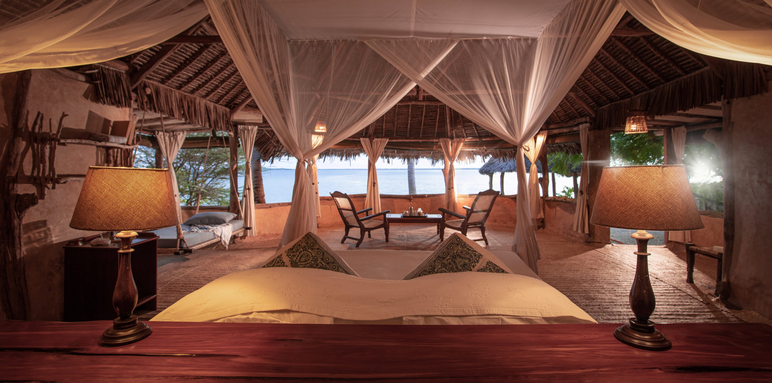 Beachfront bedroom with a mosquito-netted bed, bedside lamps, swing daybed and two planter's chairs facing open sea views, Manda Island.