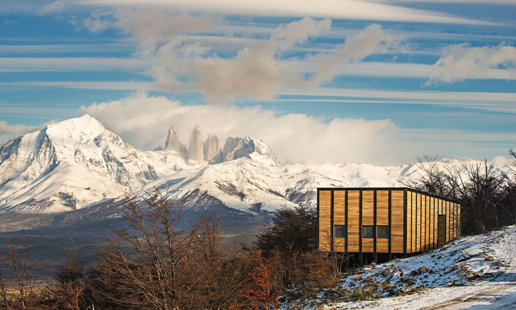 A timber-clad lodge sits on a snow-dusted Patagonian hillside, the jagged granite towers of Torres del Paine rising dramatically above snow-covered mountains behind under a sweeping sky, Chile.