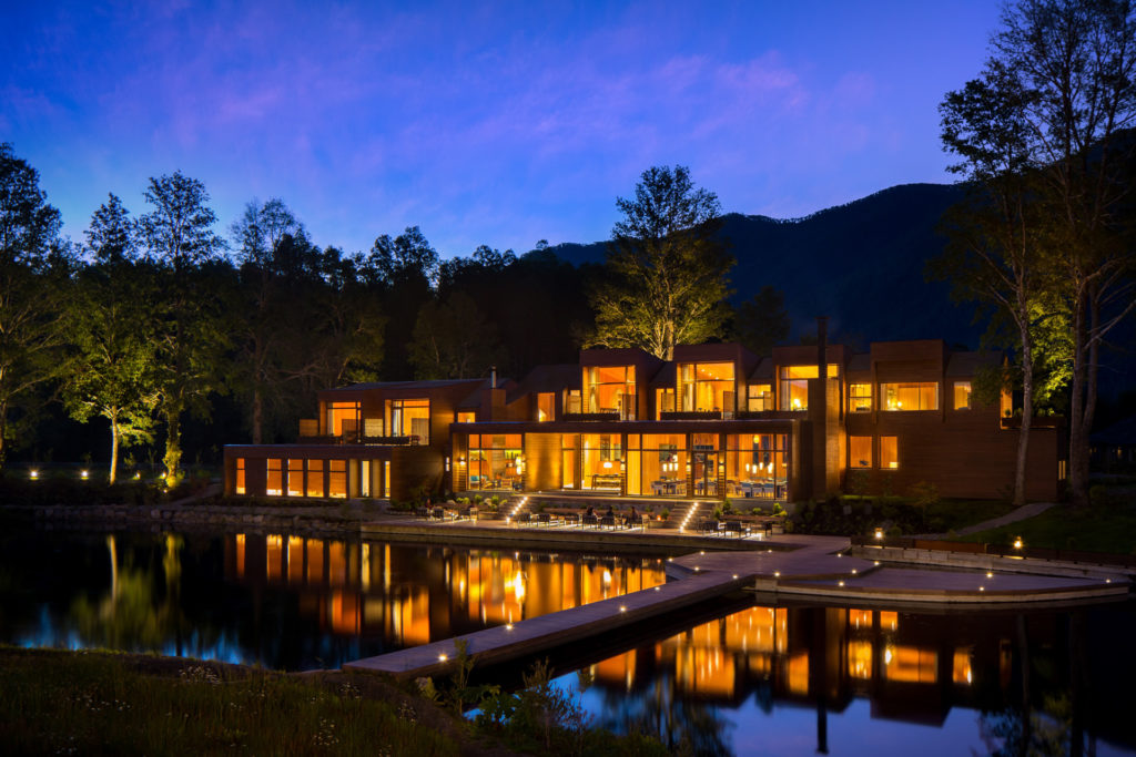 A contemporary timber lodge glows warmly with golden light at dusk, its multi-level façade reflected in a still pond in the foreground, forested mountains rising dark against a blue twilight sky, Patagonia.