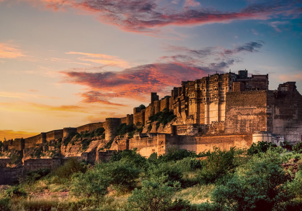 Mehrangarh Fort rising dramatically from a rocky escarpment above Jodhpur at sunset, its towering sandstone walls and ornate palace facades glowing amber and gold beneath a vivid pink and blue sky, Rajasthan, India.