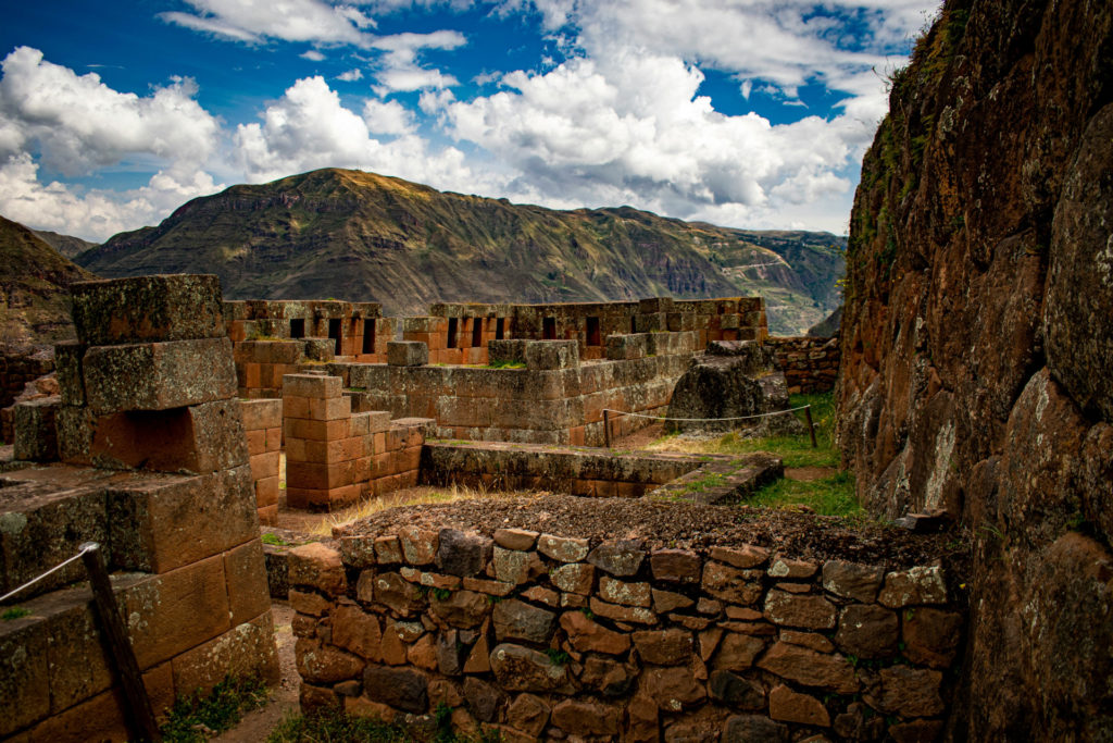 The red sandstone walls and trapezoidal niches of the Inca temple complex at Pisac, multiple roofless stone chambers stepping up the hillside beneath a vivid blue sky with white clouds and mountain ridges beyond, Sacred Valley, Peru.