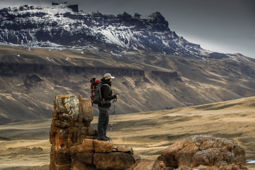 A hiker with a backpack and walking pole stands atop a lichen-covered rock formation, surveying a sweeping golden Patagonian plateau with a dramatic snow-capped mesa cliff rising in the distance under a grey sky.