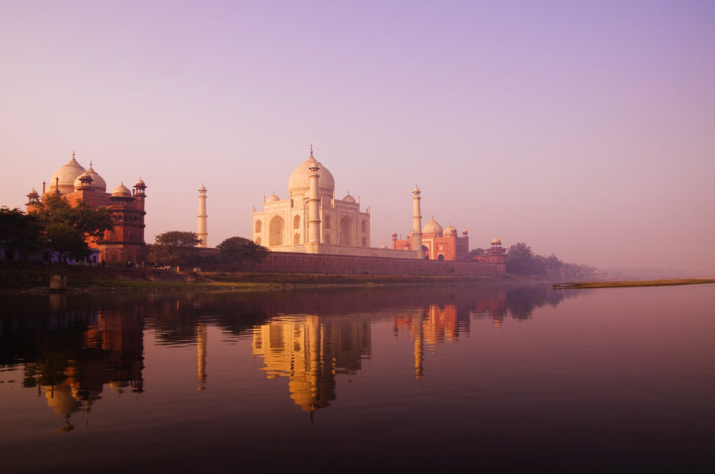 The Taj Mahal seen from across the Yamuna River at dawn, its white marble dome and minarets bathed in soft lavender and rose light, perfectly reflected in the still river below, Agra, India.