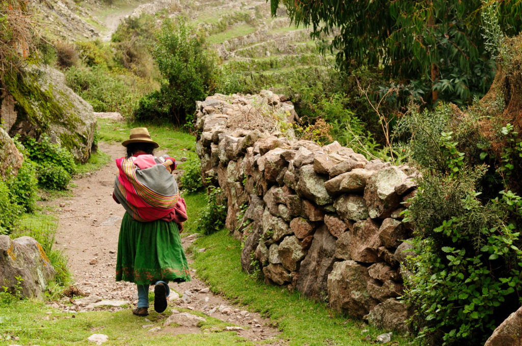 A woman in traditional Andean dress and straw hat walks along a narrow dirt path beside a dry-stone wall, a baby wrapped in a colourful cloth on her back, lush terraced hillsides beyond, Sacred Valley, Peru.