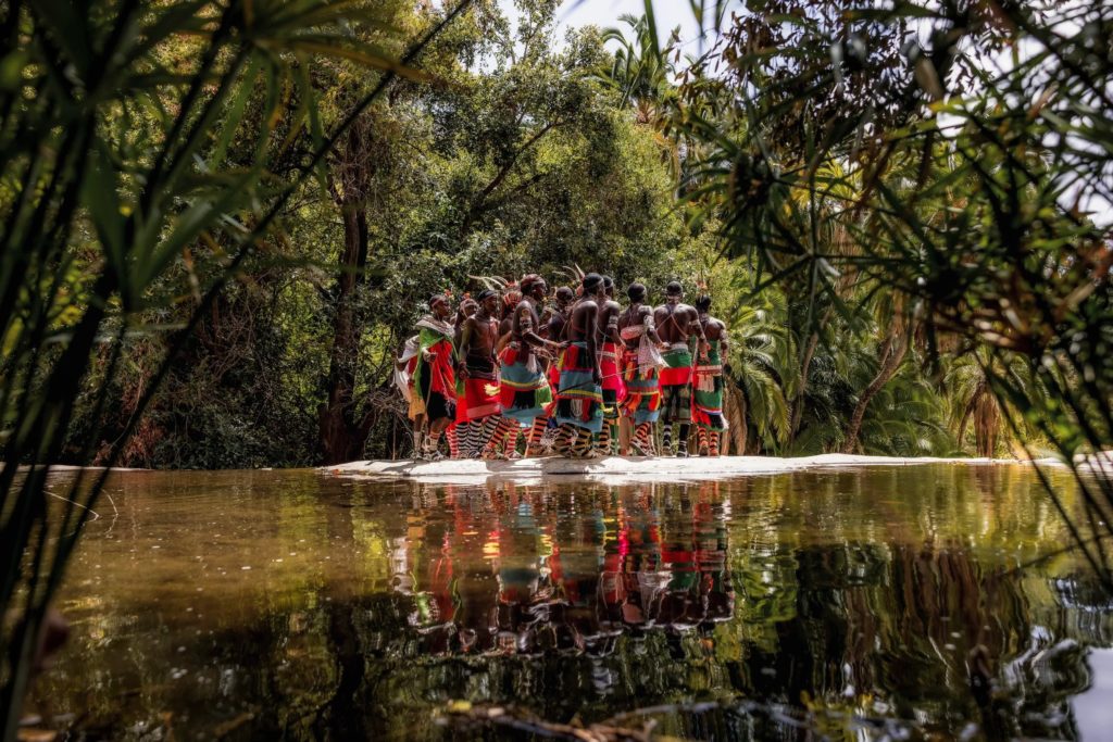 Samburu dancers in beaded collars and bright wraps gather on a rock ledge, reflected in a palm-fringed pool near Desert Rose.