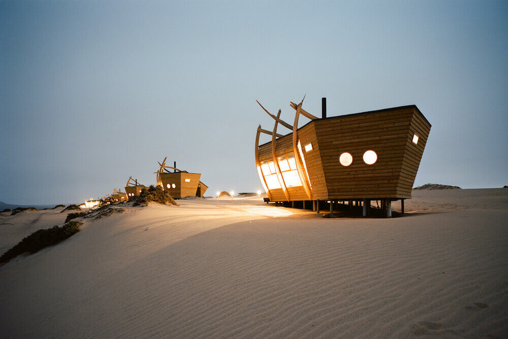 Wooden shipwreck-style chalets lit up at dusk on a Skeleton Coast dune