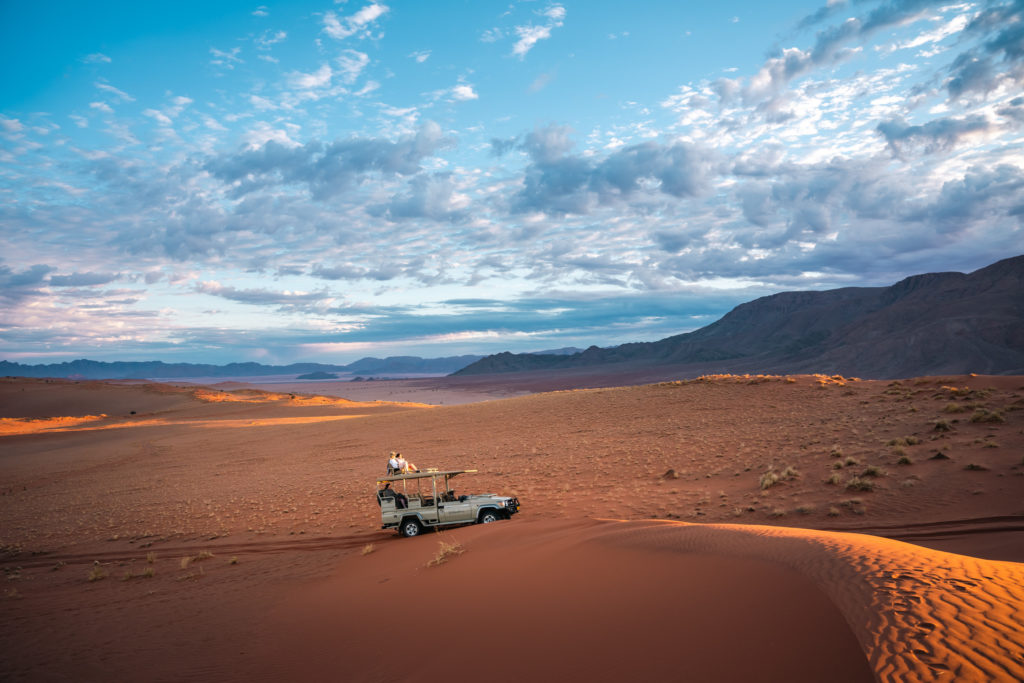 A safari vehicle parked on a red dune with guests seated on the roof at sunset