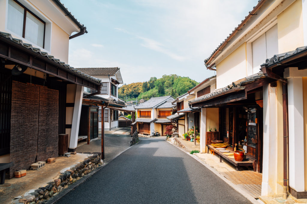 A quiet lane of old white-plastered houses with tiled roofs and wooden shopfronts, leading towards a green hill in rural Japan.