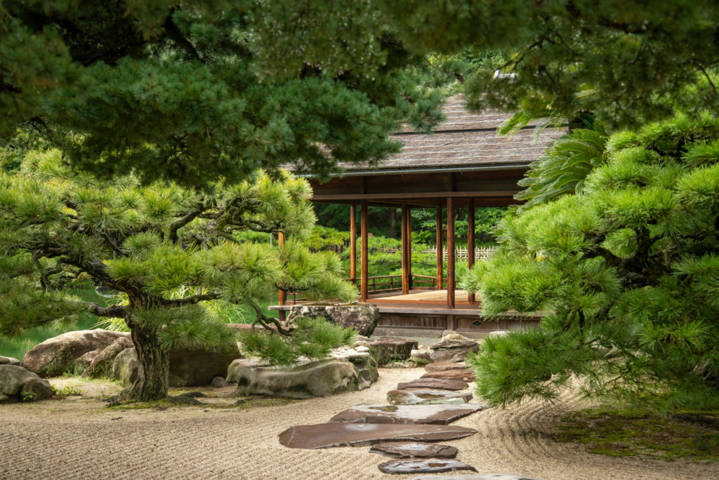 A wooden teahouse framed by pine branches in a Japanese garden, with stepping stones leading across raked gravel towards its open veranda.